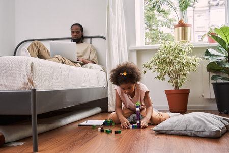 Man laying at the bed and working at the laptop while his little curly daughter sittingの写真素材