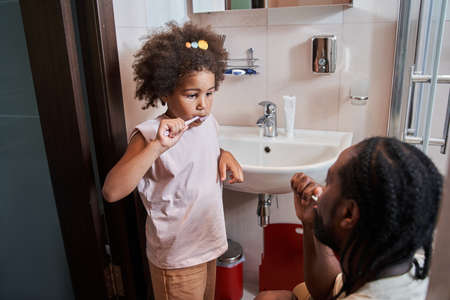 Lovely little lady cleaning teeth with toothbrush and looking at her fatherの写真素材