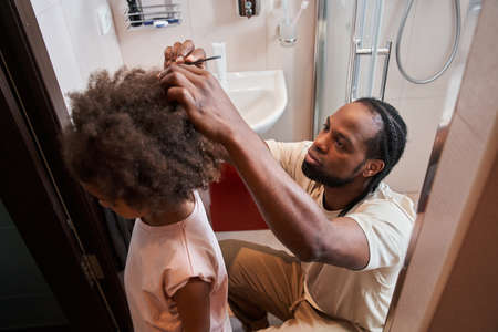 Multiracial man combing hair of his little curly daughter at the bathroomの写真素材