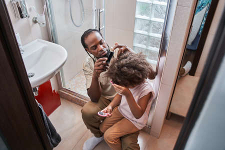 Man holding a comb and doing hairstyle for his little daughter at the bathroomの写真素材