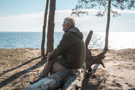 Senior man sitting at the tree log at the seashore and looking at the water surfaceの写真素材