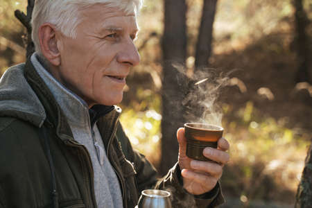 Positive tourist holding cup with hot tea while sitting on the ground in pinewoodの写真素材