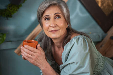 Female pensioner tasting cup of tea in living room while sitting at the stairs and looking awayの写真素材