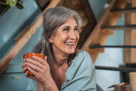 Senior woman in casual clothes sitting at the stairs and smiling while drinking hot coffee