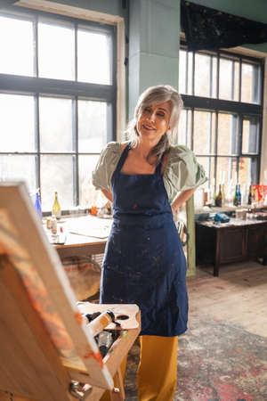 Woman ties apron on herself and looking at her masterpiece while standing in front of easelの写真素材