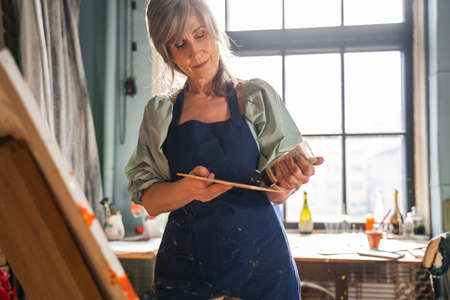 Woman in apron looking at paints while standing in front of the easel and choosing colorの写真素材