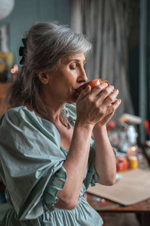 Cheerful mature woman drinking coffee while relaxing near the windowの写真素材
