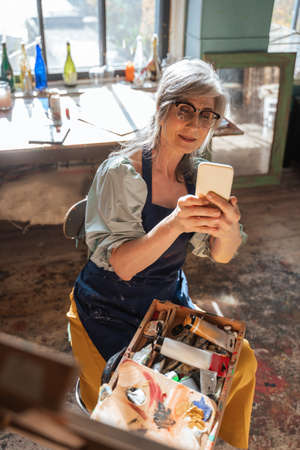 Mature artist woman sitting and shooting her masterpiece at her modern smartphoneの写真素材