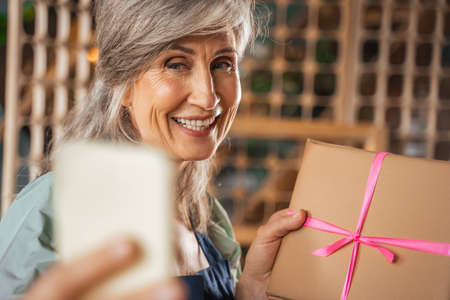 Retirement woman holding hand made gift and smiling with pleasure while taking selfieの写真素材