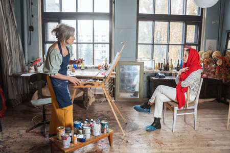 Mature female artist standing in front of the easel and looking at her model while drawingの写真素材