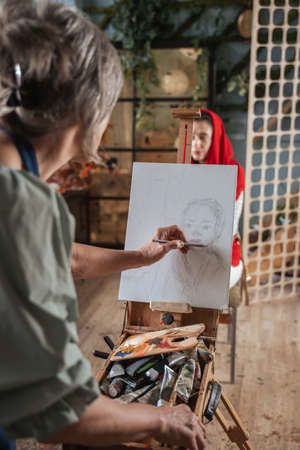 Elderly woman artist drawing with pencil on paper while standing in front of the easel