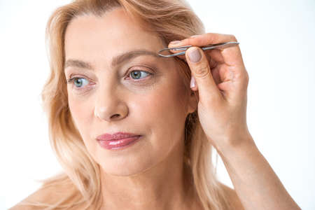 Woman correcting eyebrows with tweezers and looking awayの写真素材