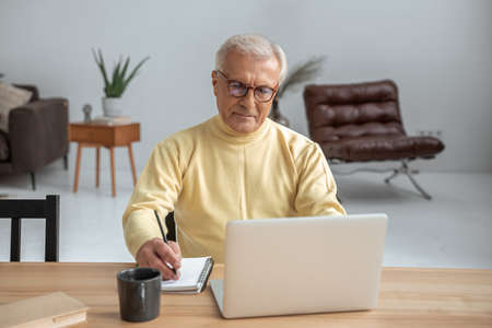 Senior man wearing glasses sitting in front of the laptop and writing notesの写真素材