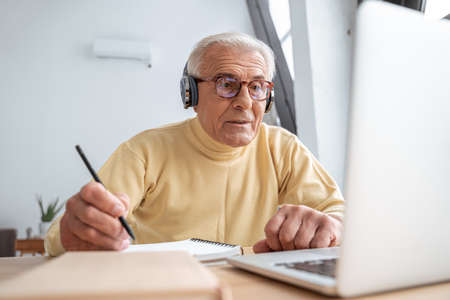 Concentrated older grandfather watching online webinar and writing notesの写真素材