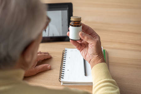 Caucasian mature man examining jar with pills while sitting at the tableの写真素材