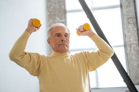 Energetic sporty mature man with gray hair exercising at home with orange dumbbellの写真素材