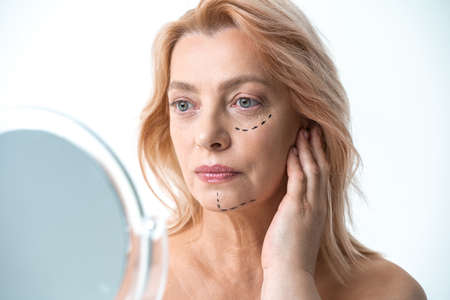 Woman looking at the mirror at her reflection while being ready for plastic surgery