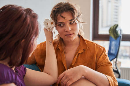 Brunette woman tucks daisies behind the ear of her girlfriend while sitting