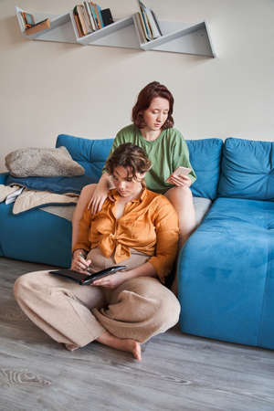 Girl sitting at sofa and browsing internet at her smartphone while her girlfriend writingの写真素材