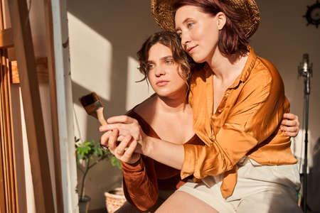 Woman wearing hat sitting at the knees of her girlfriend and drawing with her at the easelの写真素材