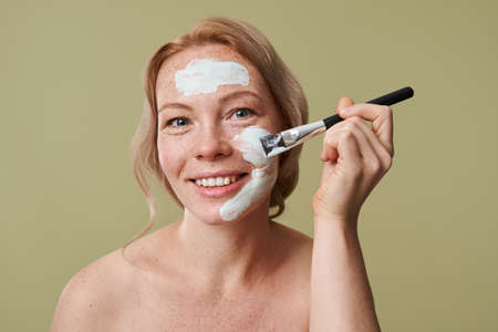 Woman applying clay mask wit brush, looking at camera isolatedの写真素材