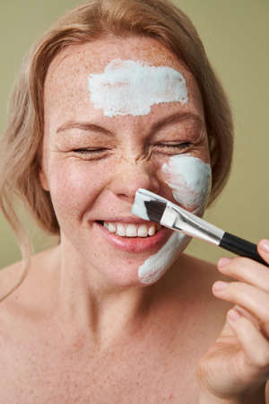 Beautiful girl applying facial clay mask and laughing out loud isolatedの写真素材