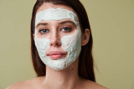 Brunette young woman with clay facial mask looking at the camera seriouslyの写真素材