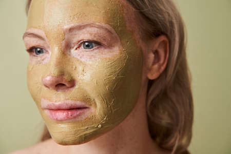Attractive girl with clay mask on face looking away isolated over green backgroundの写真素材