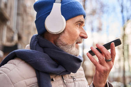 Senior guy wearing headphones recording voice message at his smartphoneの写真素材
