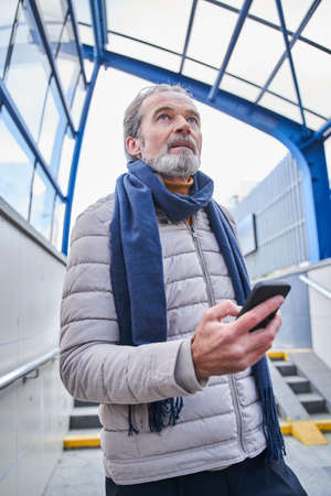 Concentrated elderly male standing outdoors and holding mobile