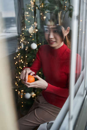 Asian woman sitting at the windowsill and peels tangerineの写真素材