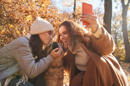 Overjoyed teenage girl kissing her lovely dog while mother making selfie with her family