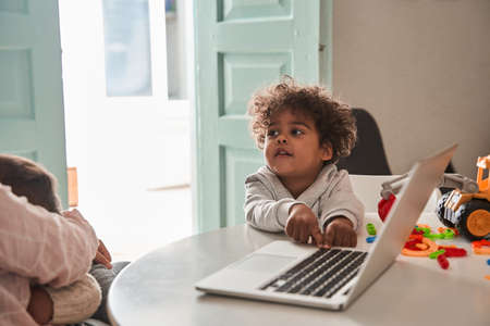 Curly multiracial curious boy touching keyboard of the laptop while sitting at the tableの写真素材