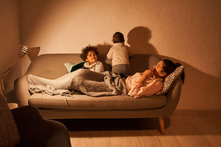 Kids spending time at sofa with their mother while laying together and playing during the eveningの写真素材