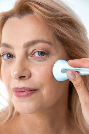 Mature woman with clean face skin holding massager in her hand while taking care of skinの写真素材