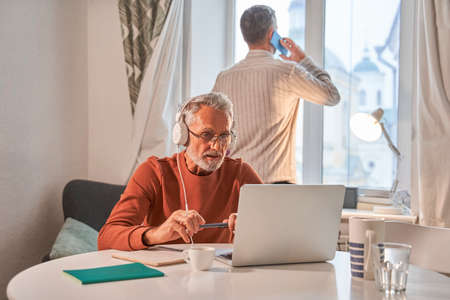 Adult man talking via smartphone while his father having video call at the laptopの写真素材