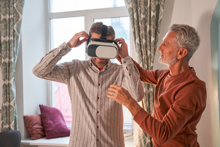 Bearded senior man helping to his son wearing glasses of virtual reality while standingの写真素材