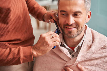 Handsome man getting his beard cut at home while his careful father helping to him