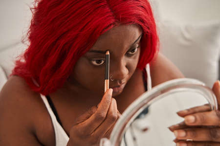 Multiracial woman with red hair making make up while sitting at the sofa and looking at the mirrorの写真素材