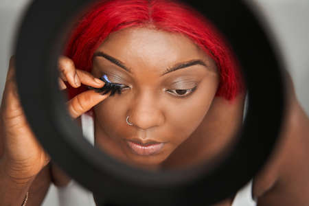 Calm multiracial woman putting false eyelashes at her face attentively while sitting at home