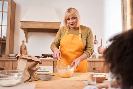 Attentive woman looking at her female child while mixing ingredients with whisk into the bowl