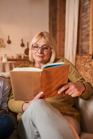 Senior woman holding book and feeling concentrated while reading at the sofaの写真素材