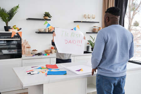 Boy showing placard to his daddy while congratulating him with Fathers Day at the kitchenの写真素材