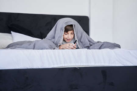 Boy hiding under the blanket while laying at the bed and playing with walkie-talkie at homeの写真素材