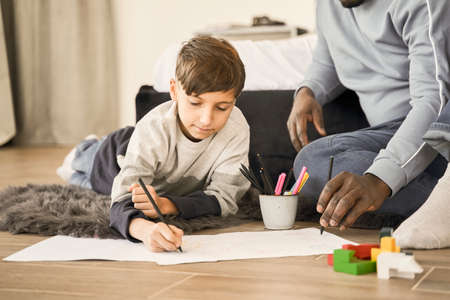 Multiracial man sitting near his caucasian son and looking at him while drawing togetherの写真素材