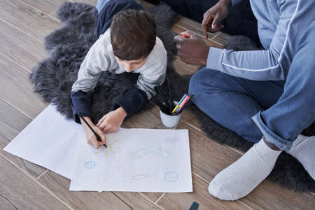 Boy drawing hand made picture with his father while sitting at the floor at cozy workplaceの写真素材