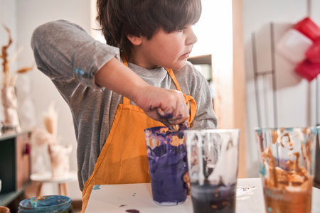 Boy standing with serious face and putting his hands into the plastic cup with violet paintsの写真素材