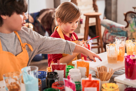 Boy pointing with his finger at the glass with paints while drawing near his brother or friendの写真素材