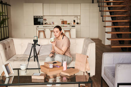 Female blogger influencer sitting at home and looking at cream while shooting herself on smartphoneの写真素材