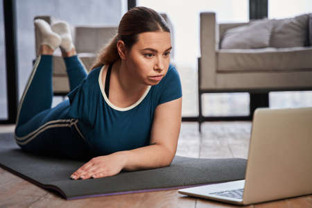Caucasian woman with amputee arm laying at the yoga mat and watching something at the laptop